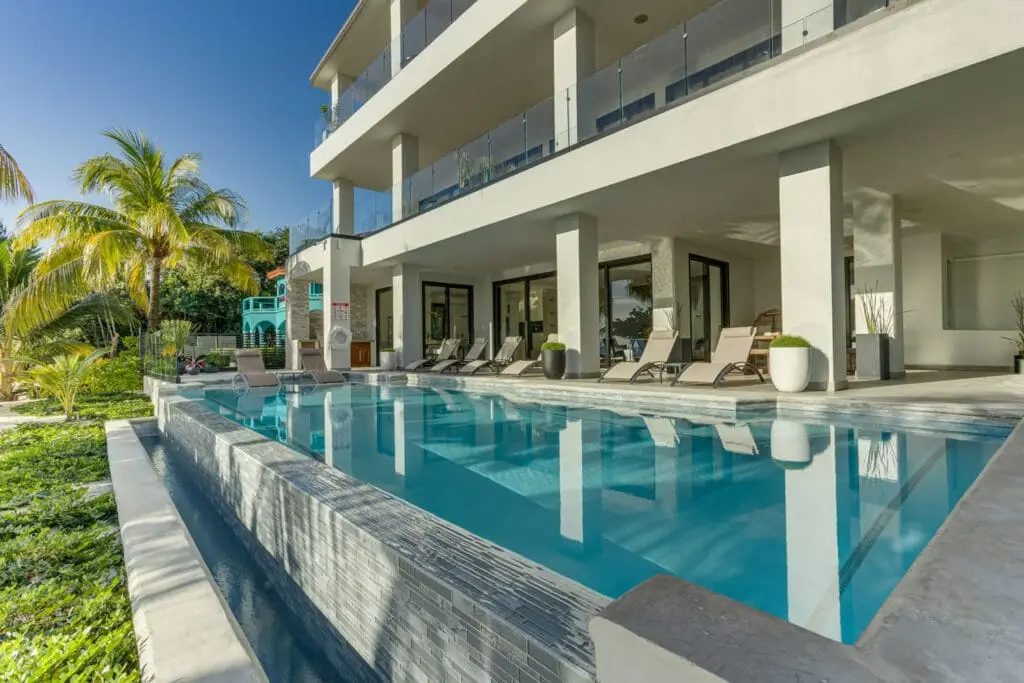 A luxury white beachfront villa with a large, clear private swimming pool, sun loungers, and palm trees under a bright blue sky.
