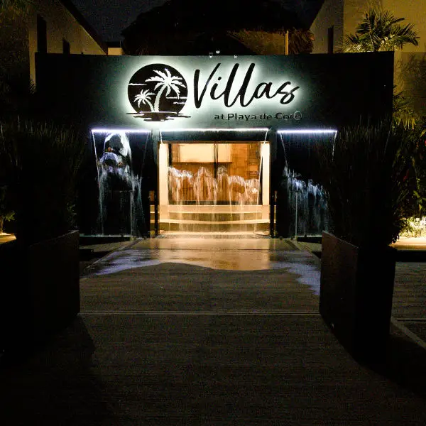 The illuminated entrance of Villas at Playa de Coco at night, featuring a lit resort sign and a walk-through waterfall feature.