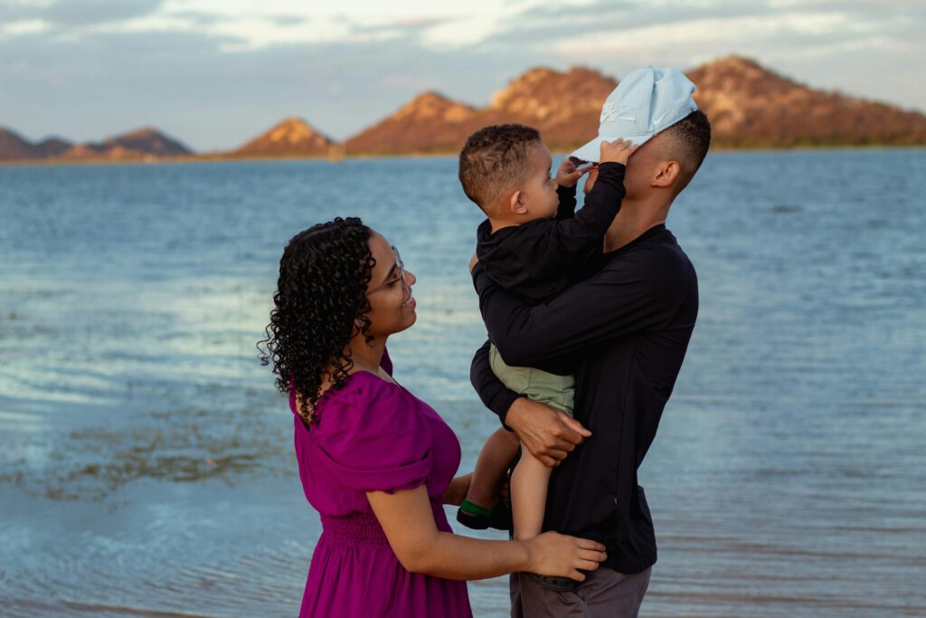 A happy family of three on vacation, with a father lifting his young son into the air while the mother looks on smiling, set against a scenic backdrop.