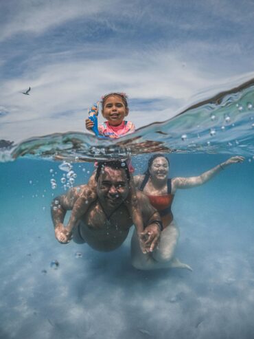 A family of four happily snorkeling and swimming together underwater in clear blue tropical water.