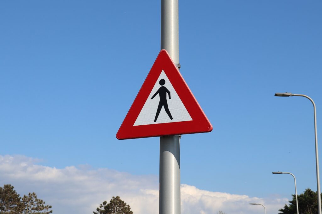 A triangular traffic warning sign with a red border showing a black silhouette of a person walking, set against a clear blue sky.