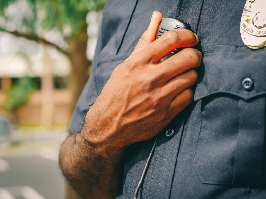 A close-up photograph focusing on the hand and chest of a male security officer wearing a dark blue uniform. His left hand is resting on his chest near the shoulder, and a portion of his 'PRIVATE SECURITY' badge is visible.