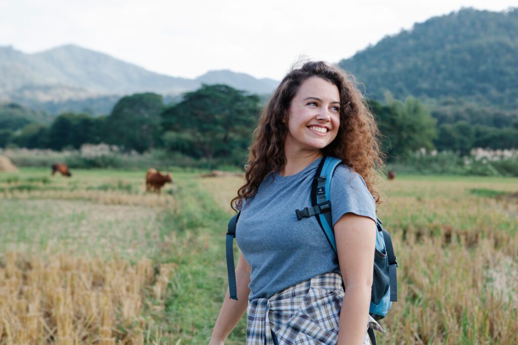 A smiling young woman with curly hair looks back over her shoulder while wearing a blue backpack on a trail with green hills in the background.